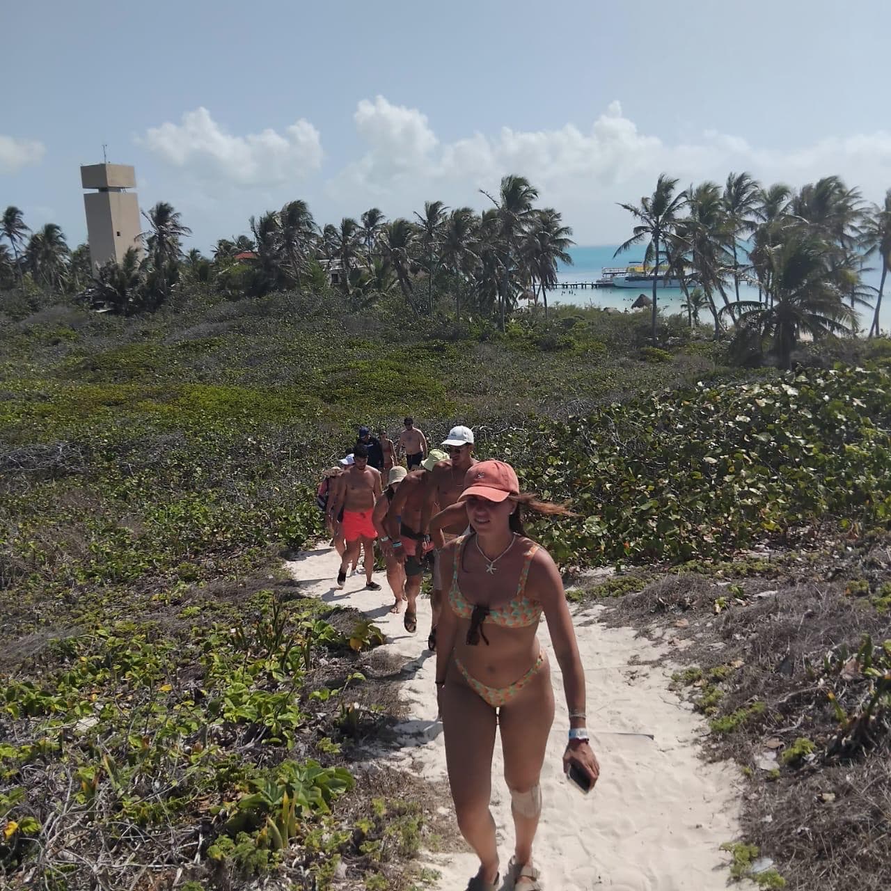 Huéspedes caminan por un sendero de arena entre vegetación costera rumbo al muelle